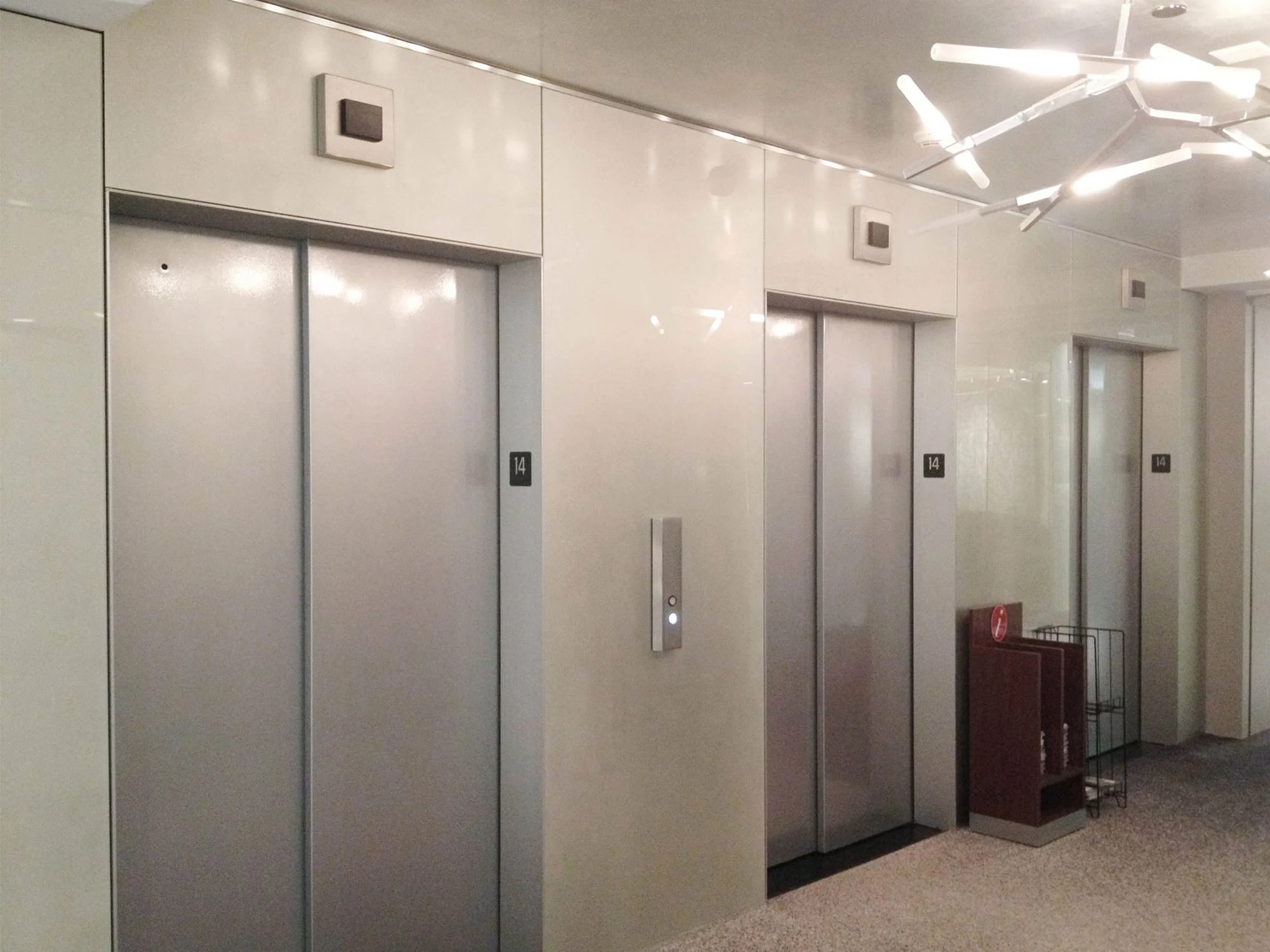 백페인트글라스_엘리베이터_실버_로비벽면_시공 현대적인 조명과 글로시한 실버 색상의 백페인트글라스로 마감된 오피스 건물의 엘리베이터 홀 벽면